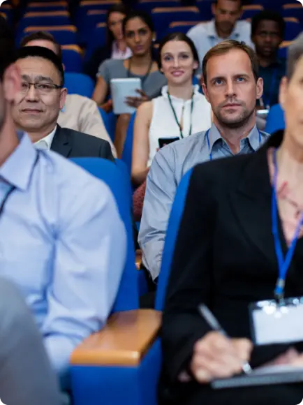 Diverse group of business professionals seated in blue auditorium chairs listening to a presentation